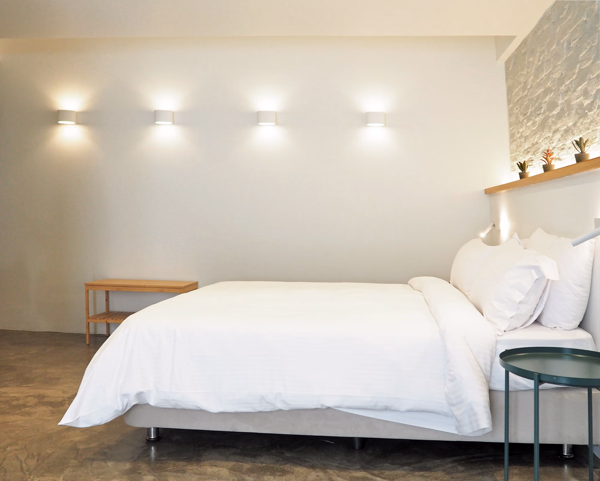 Minimalist bedroom with white bed, wooden bench, green side table, and wall-mounted lights.