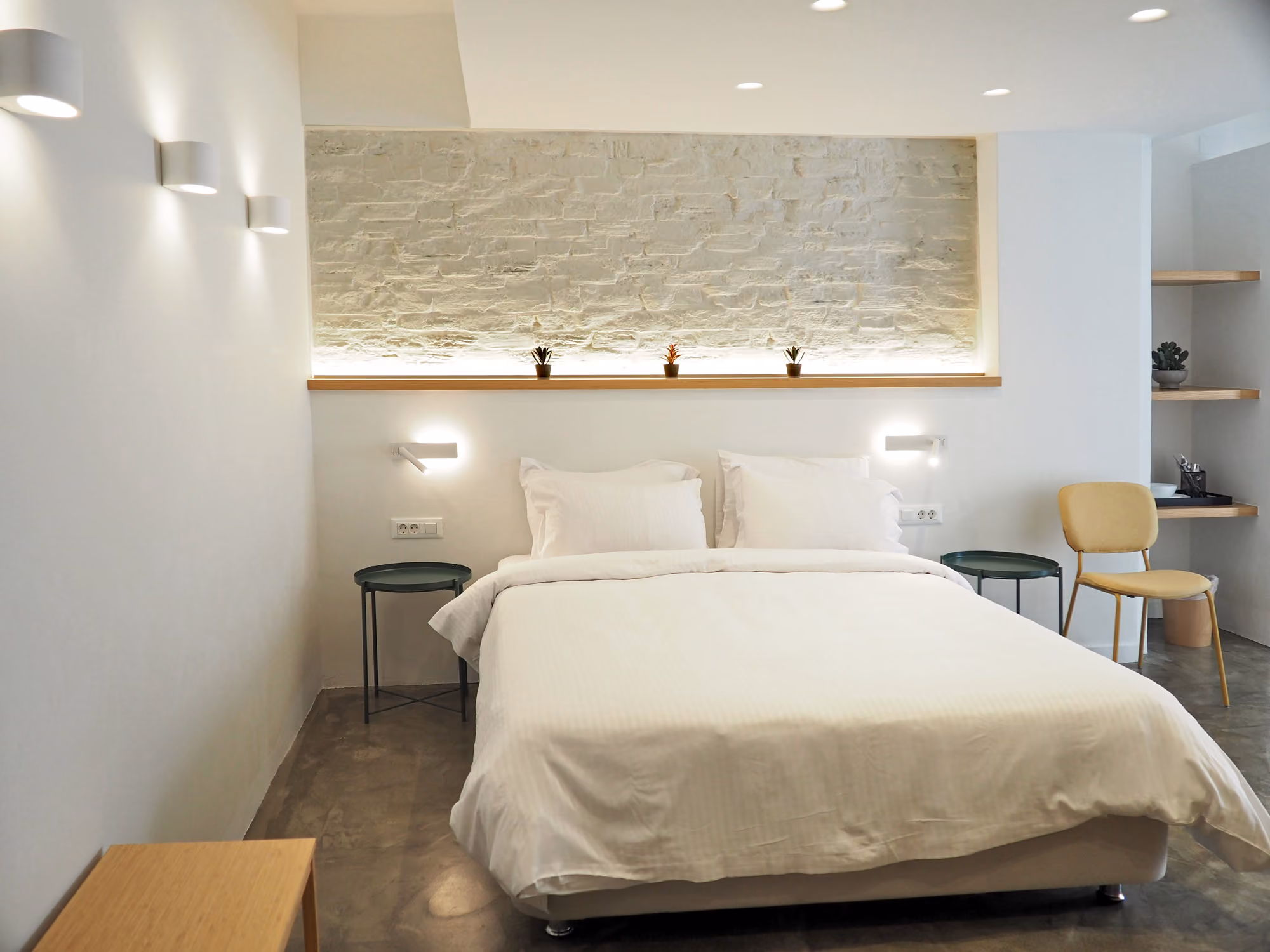 Minimalist bedroom with white bed, stone accent wall, small plants, two side tables, and a yellow chair.