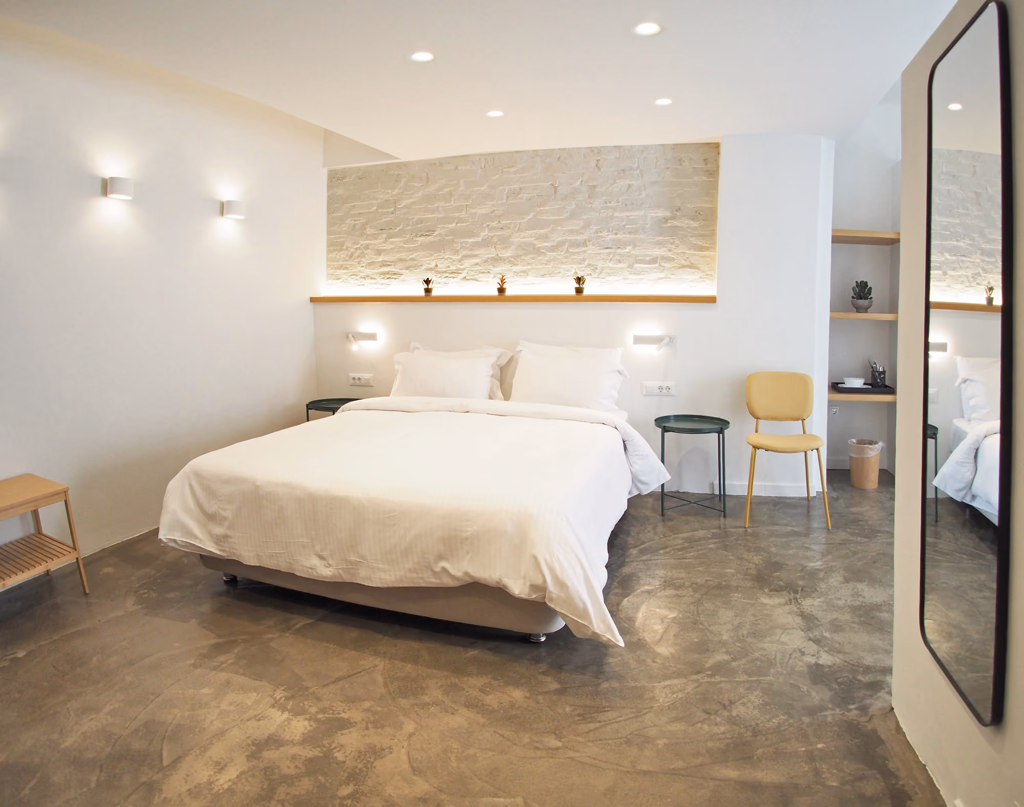 Minimalist bedroom with white bed, two side tables, yellow chair, concrete floor, and a wall mirror.