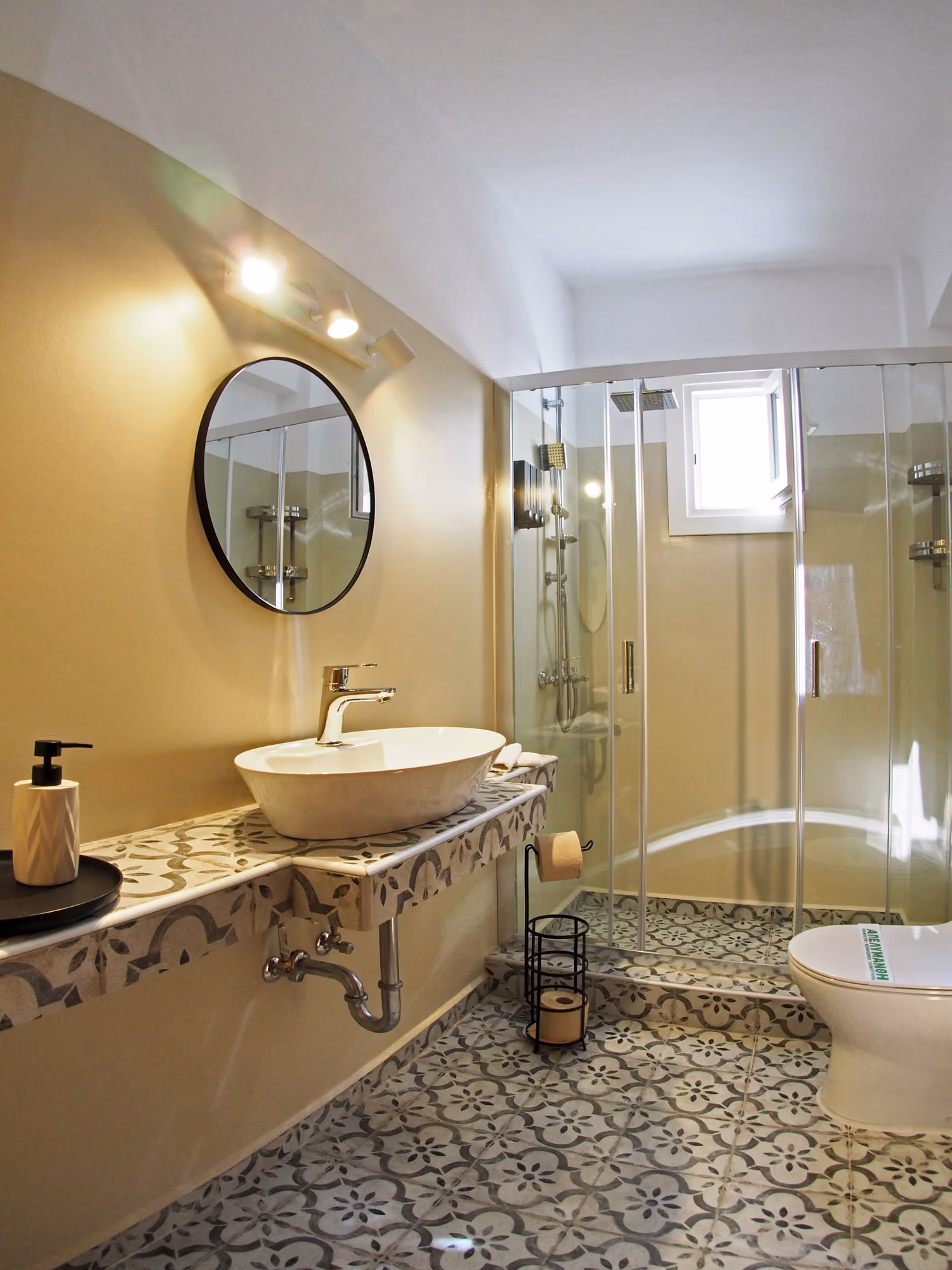 Modern bathroom with patterned floor tiles, round mirror, vessel sink, glass shower, and toilet.