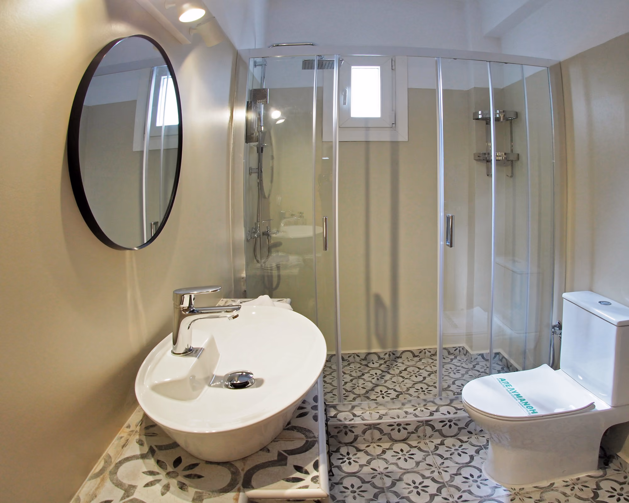 Modern bathroom with oval sink, round mirror, patterned floor tiles, glass shower, and white toilet.